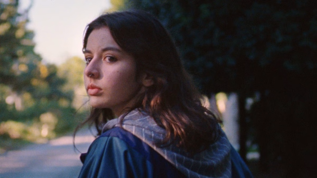 Day-in-the-life-of-jo-chapter-phaedra_nyff64_01jpg | the Cinema Group Young Woman with Brown Hair in a Blue Jacket Looking over Her Shoulder on a Sunlit Street Outside Park Trees.