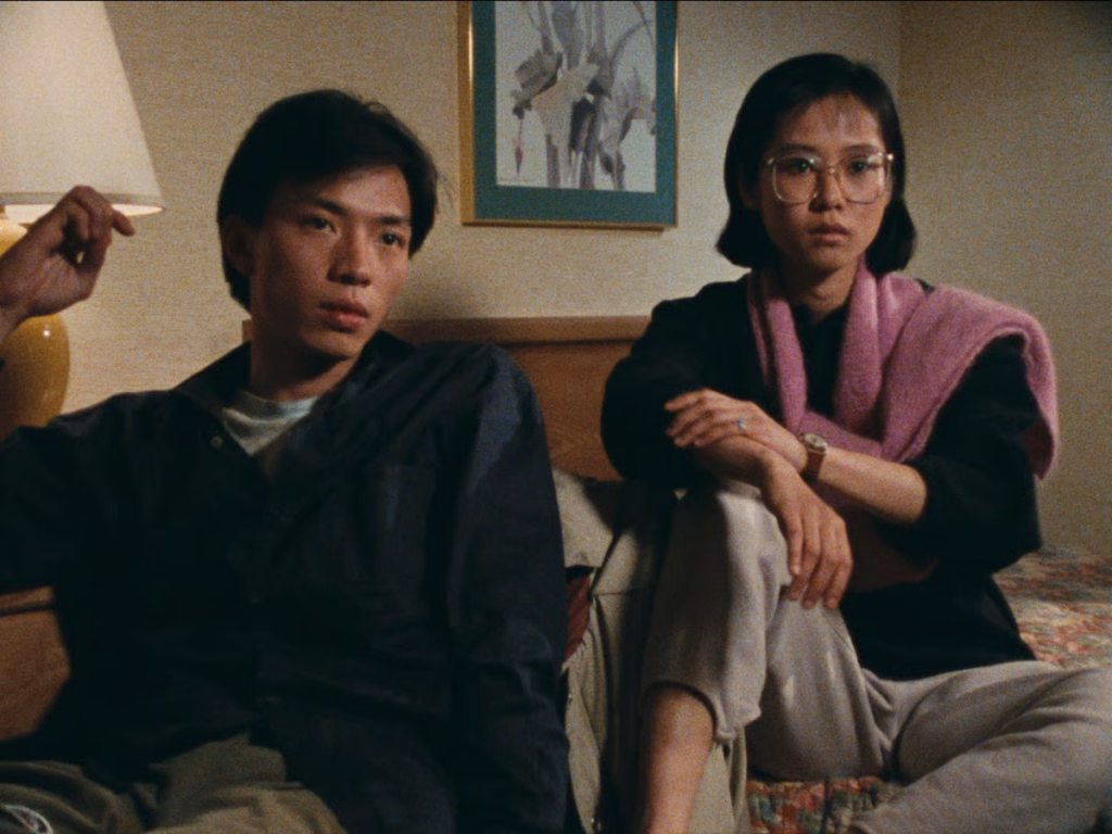 Just-like-weather_nyff64_02-2jpg | the Cinema Group Two People Sit on a Bed in a Dim Hotel Room, Looking Pensively at Something Off-screen, Neutral Expressions.