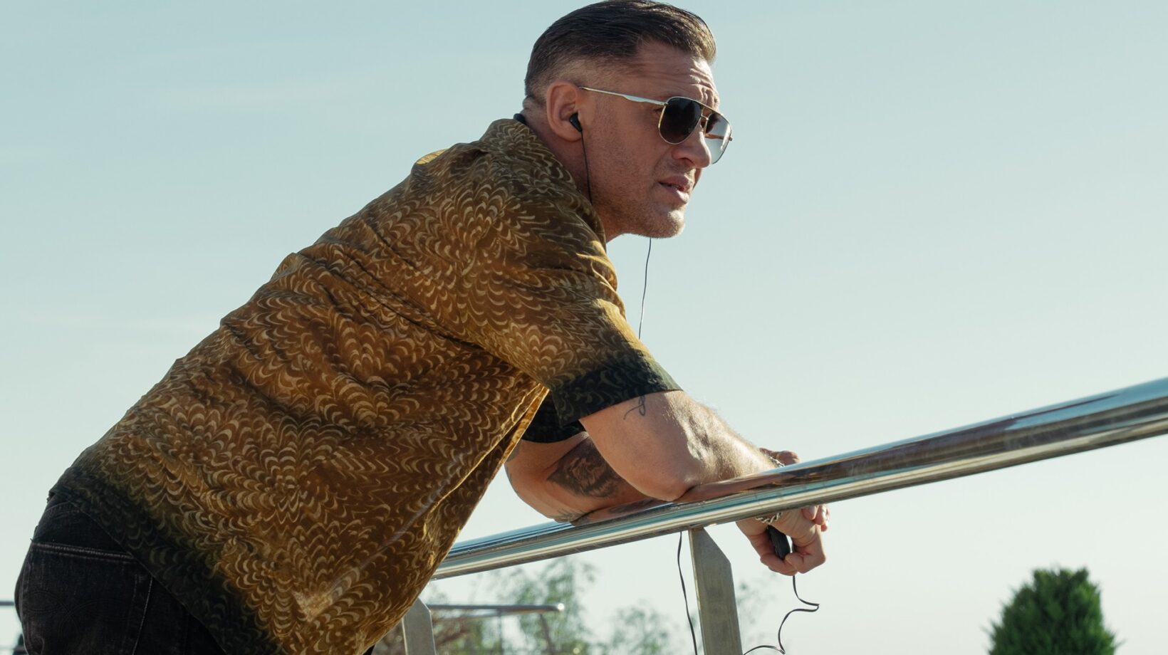 Tom Hardy Leaning over a Metal Railing, Wearing a Brown Patterned Jacket and Sunglasses, Listening to Earphones Outdoors in Pararmount+'s Mobland.