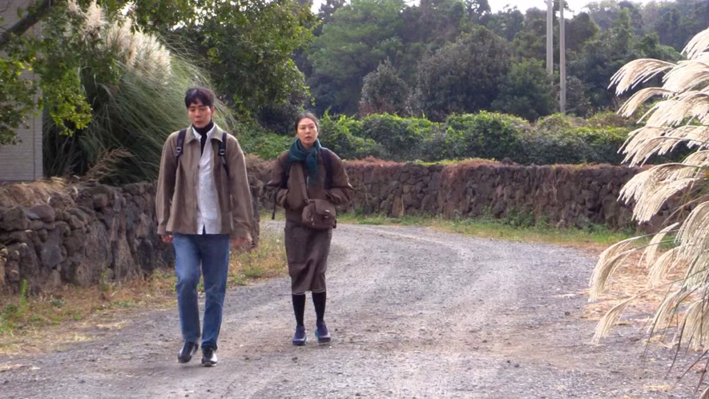 Nowhere-to-lay-my-eyes_nyff64_01jpg | the Cinema Group Two People Walk Along a Gravel Rural Path Lined with Stone Walls and Greenery on a Sunny Day.