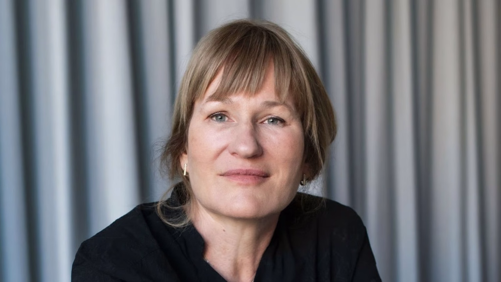 Talks_valeska-grisebachpng | the Cinema Group Portrait of a Middle-aged Woman with Blonde Hair, Wearing a Black Top, Looking at the Camera in Front of Draped Curtains.