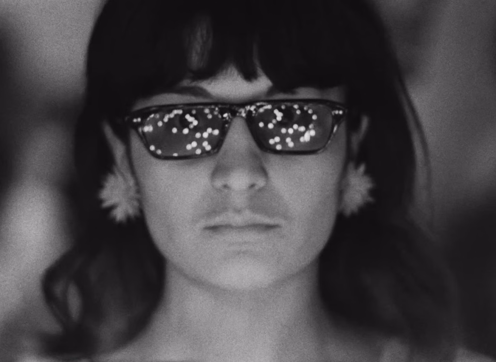 We_nyff64_01-e1786721286561-2png | the Cinema Group Woman Facing Camera Wearing Dark Sunglasses Reflecting Lights, with Flowers in Her Ears in a Black-and-white Portrait of a Calm Mood