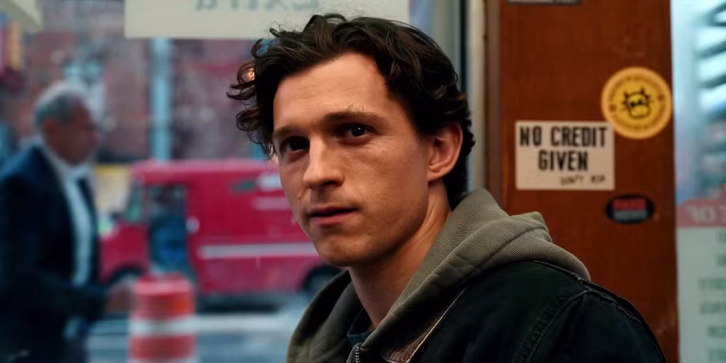 Spider-man-brand-new-day-tom-holland-bodega | the Cinema Group Young Man with Dark, Wavy Hair Wearing a Gray Hoodie and Black Jacket, Looking Toward the Camera Inside a Doorway; Blurred City Street Visible Through the Glass Behind Him.