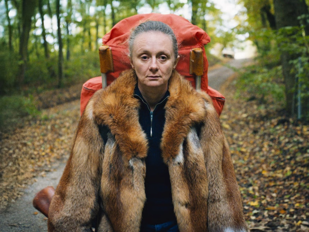 Tomorrow-a-long-time-ago-stills_nyff64_02jpeg | the Cinema Group Woman with a Large Fur Coat and Red Backpack Walking Down a Forest Path in Autumn Leaves, Looking Straight at the Camera.