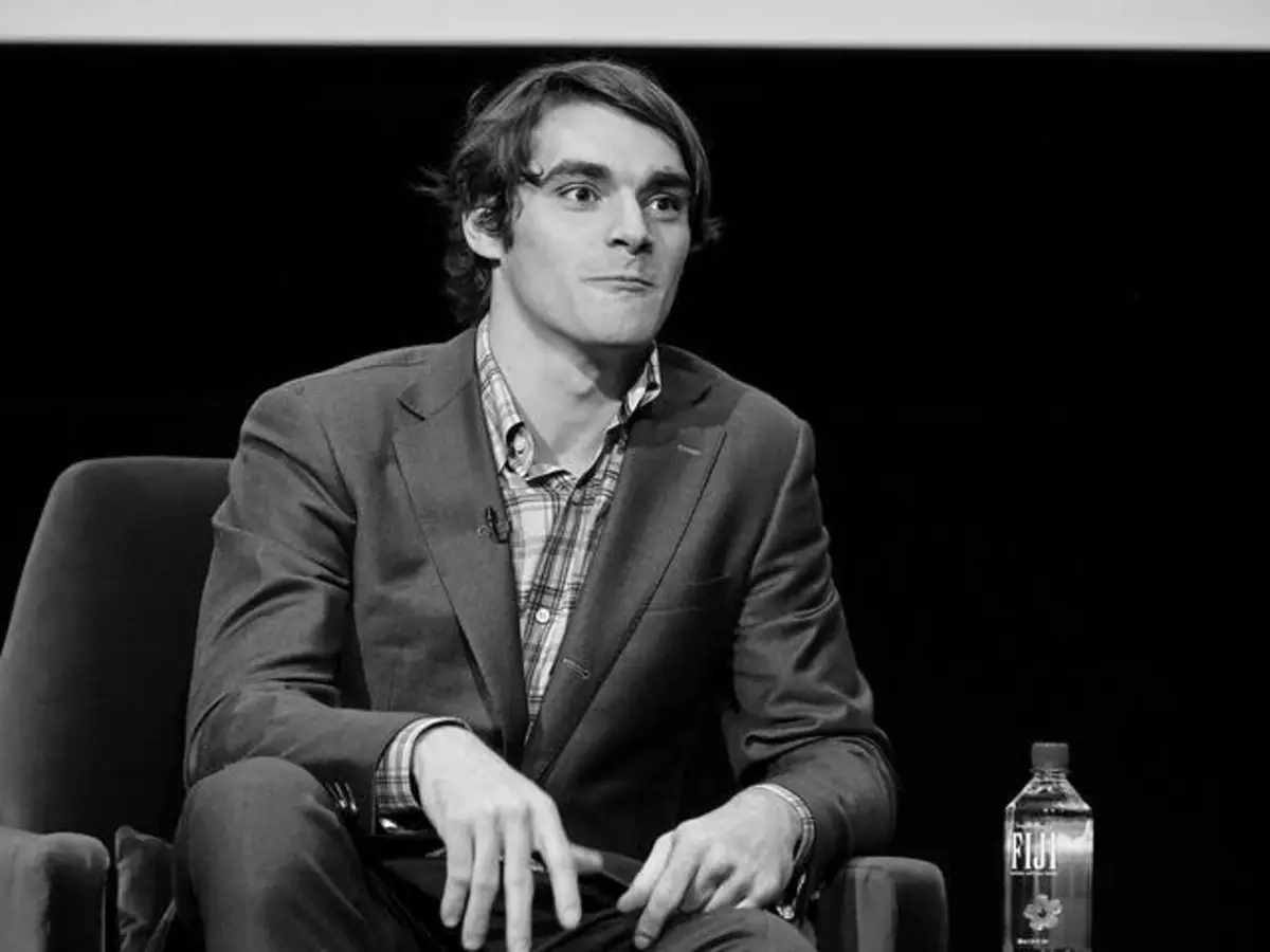 Black-and-white Photo of a Man in a Blazer and Plaid Shirt Seated on a Stage, Hands Resting on His Lap, with a Water Bottle on a Table Nearby.