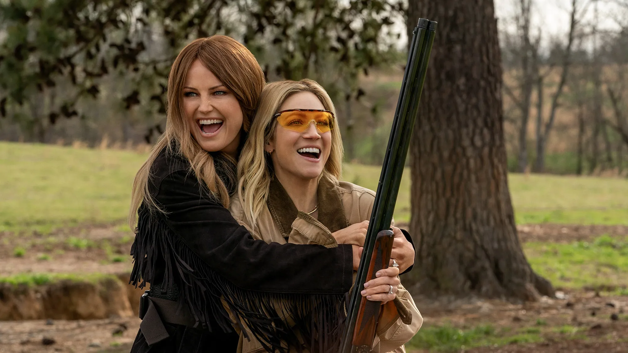 Two Women Smile and Hug Outdoors While One Holds a Longgun, Wearing Safety Glasses, in a Park Setting.
