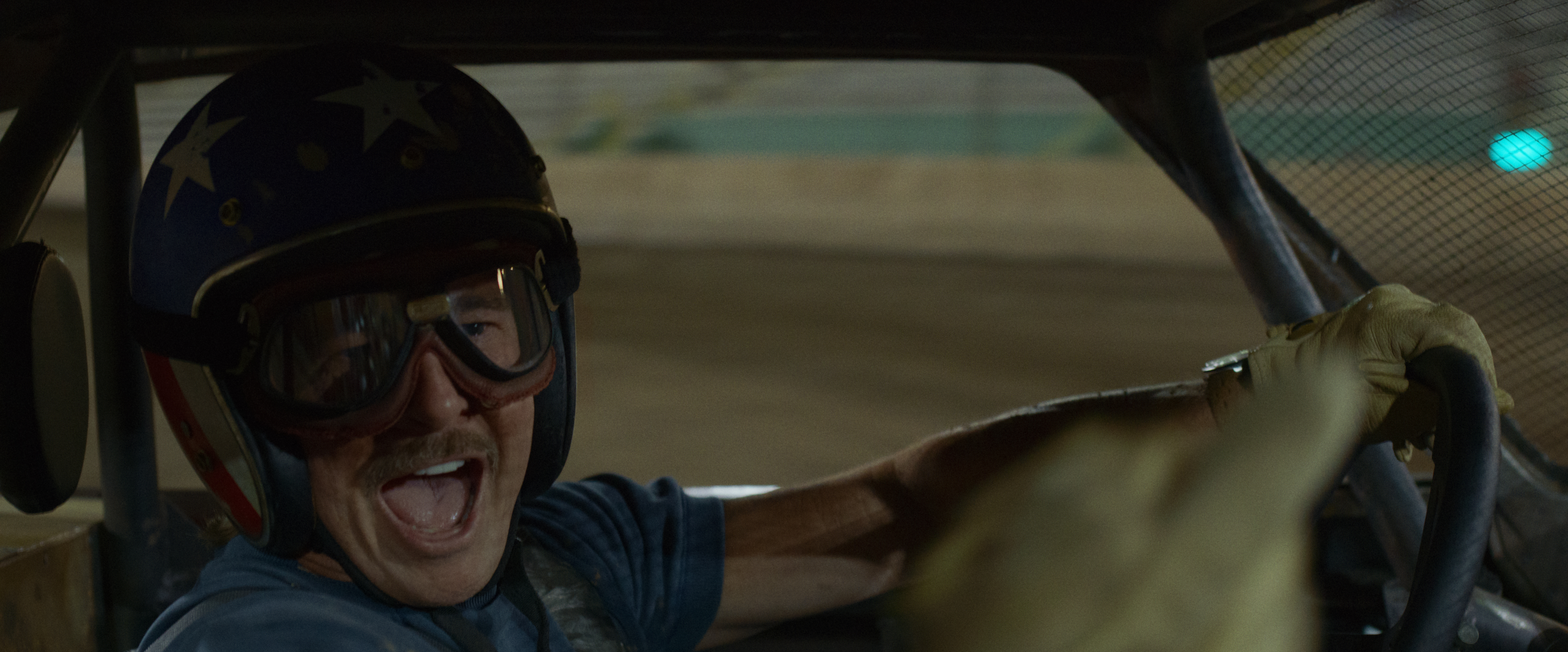 the Further Mis-adventures of Cliff Booth | the Cinema Group "man Wearing a Helmet and Goggles Smiles Excitedly While Driving a Race Car on a Dirt Track, Visible Through the Vehicle’s Interior, Suggesting a Lively Racing Atmosphere."