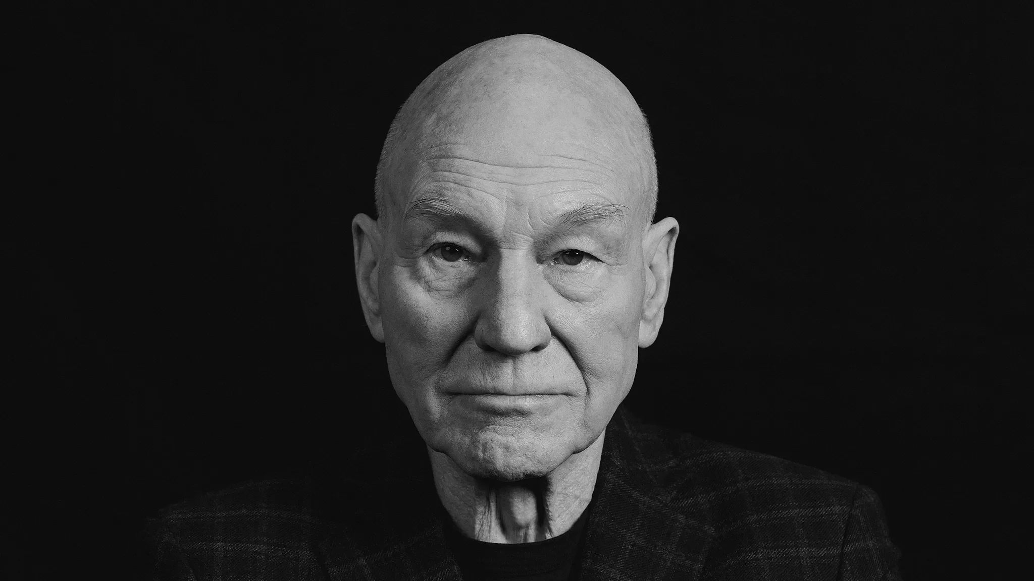 Close-up Portrait of an Elderly Bald Man with a Calm Expression Against a Solid Black Background, Wearing a Dark Textured Blazer.