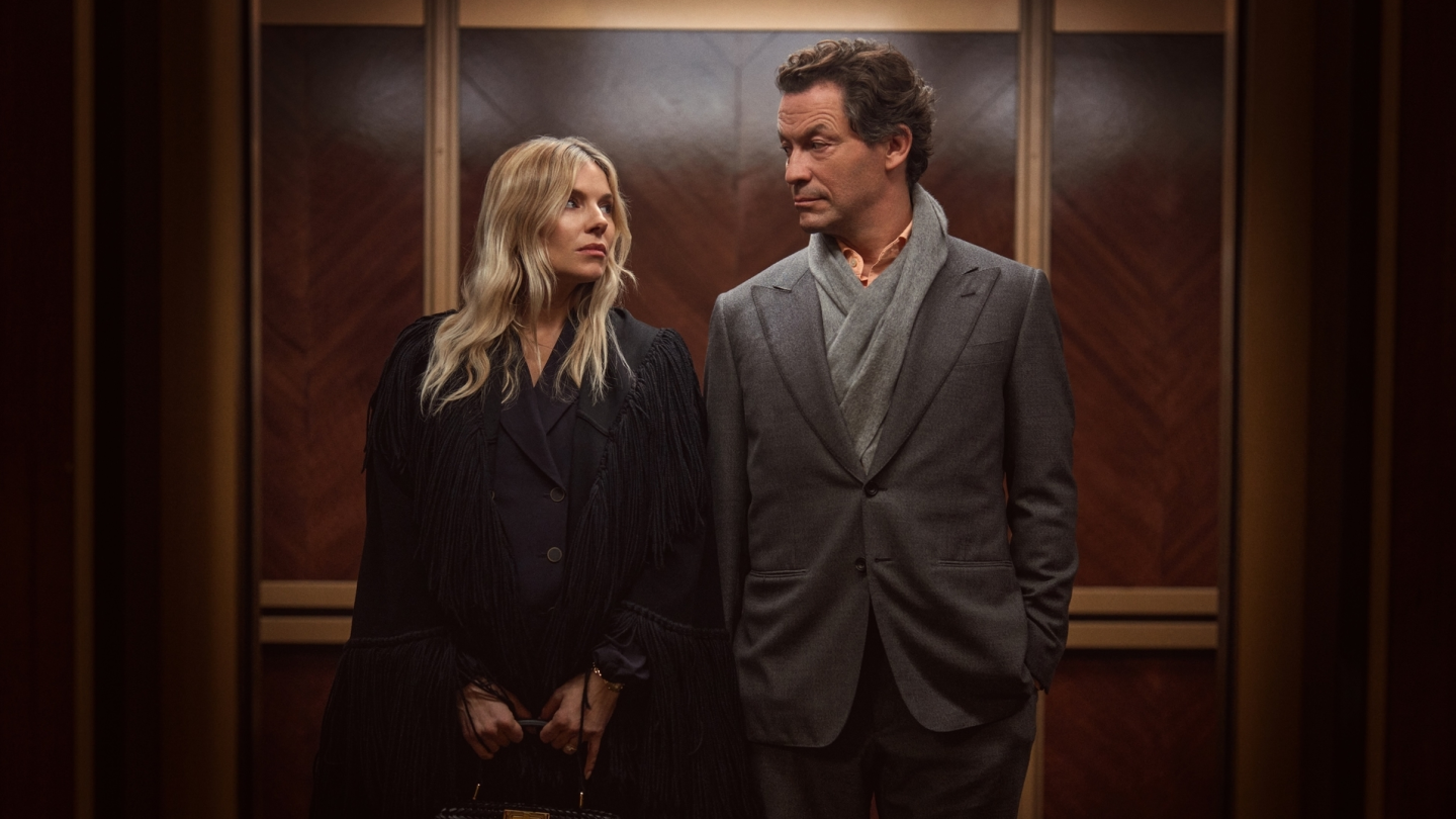 a Man in a Gray Suit with a Light Scarf Stands Beside a Woman in a Black Fringed Coat Inside a Wood-paneled Elevator Lobby, Both Looking at Each Other.